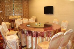 Mesa redonda com tampo de vidro, rodeada por cadeiras com capas estampadas, televisão de tela plana na parede e cortina decorativa ao fundo.