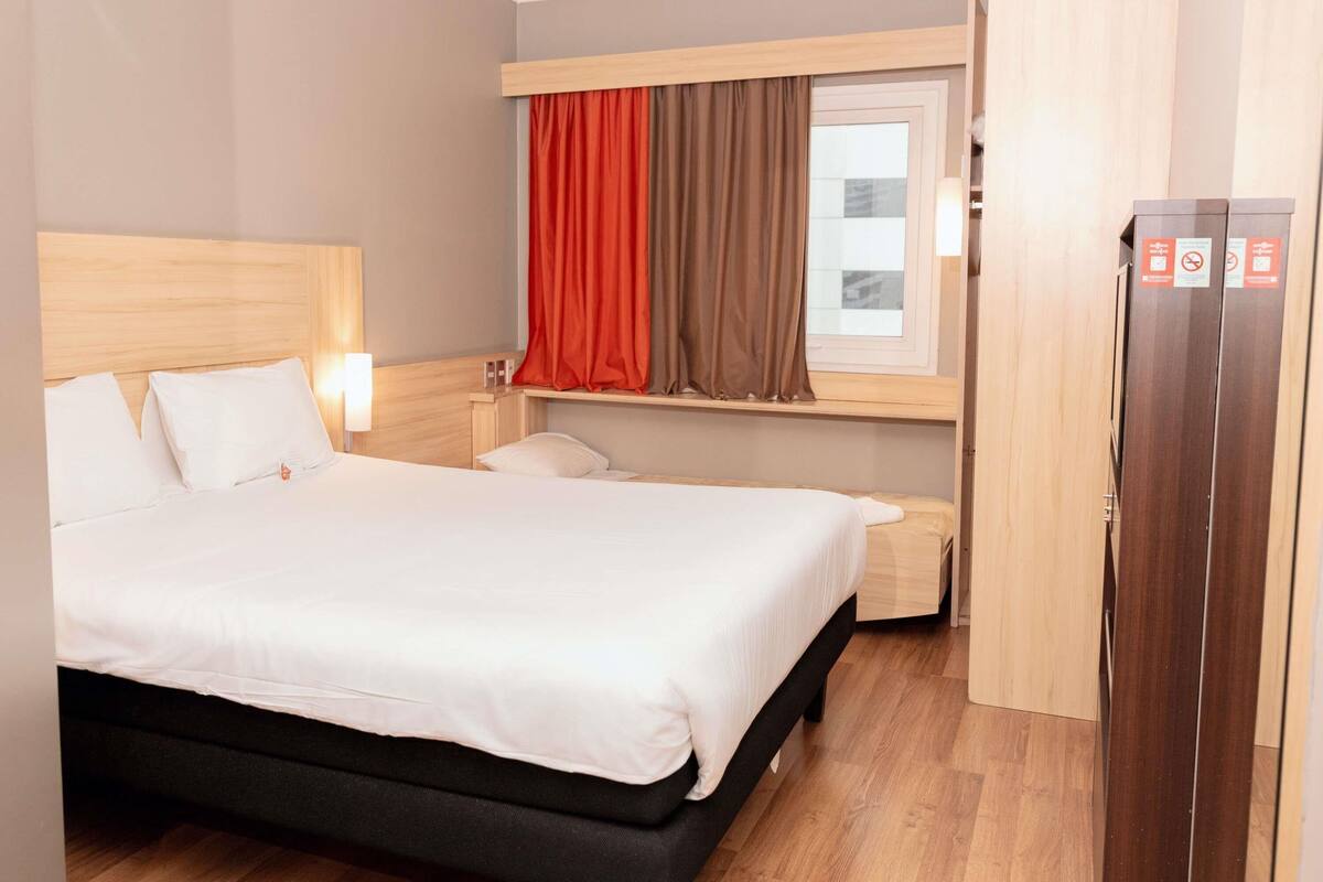 A hotel room with a large bed, a wooden headboard, a window with curtains, a bedside table, and a wardrobe.