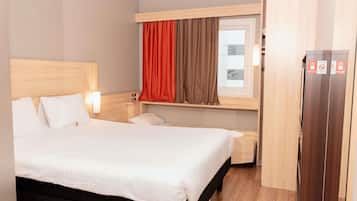 A hotel room with a large bed, a wooden headboard, a window with curtains, a bedside table, and a wardrobe.