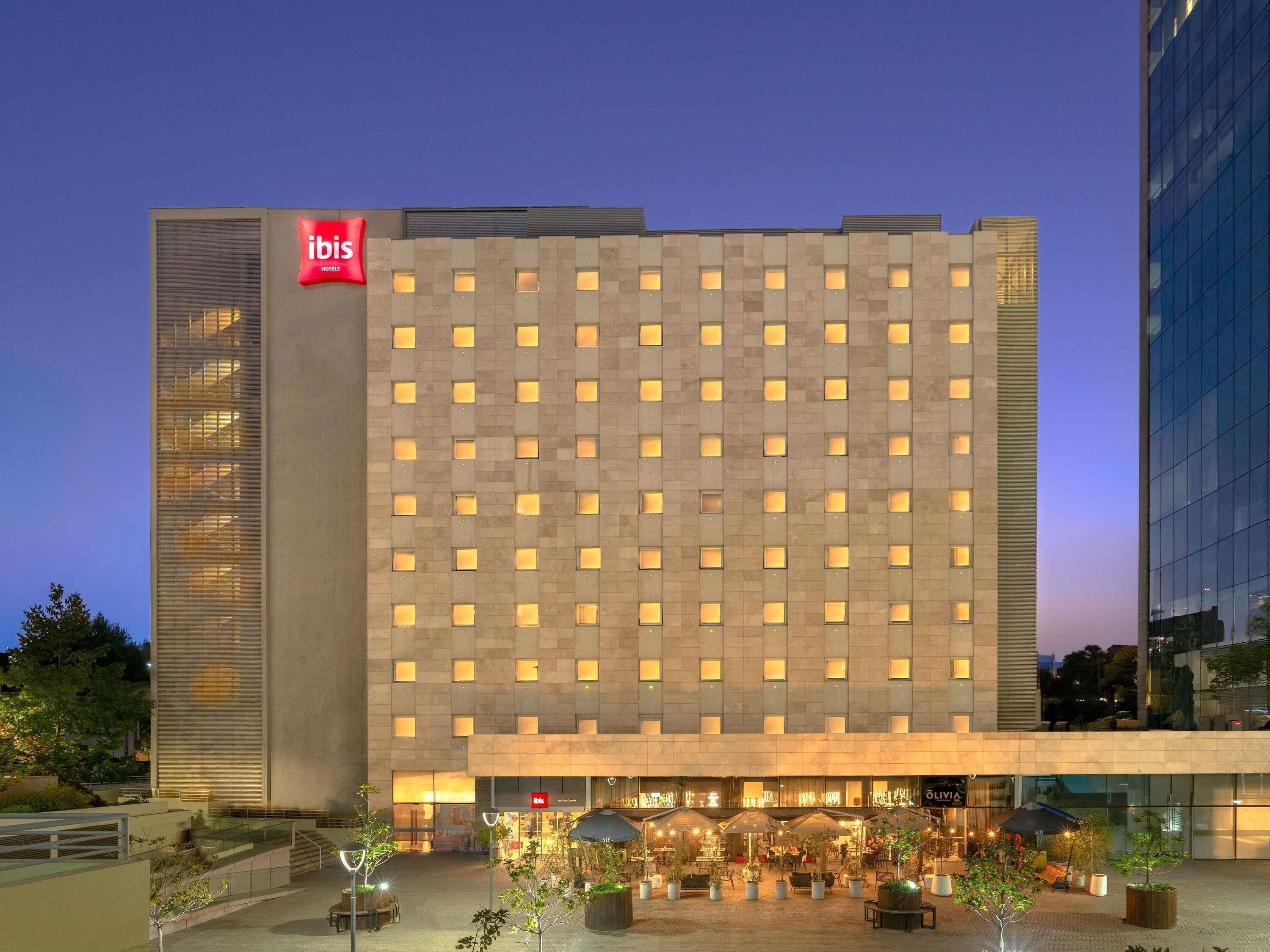 A modern hotel building with a prominent 'ibis' sign, a courtyard with outdoor seating, and a glass facade on one side.