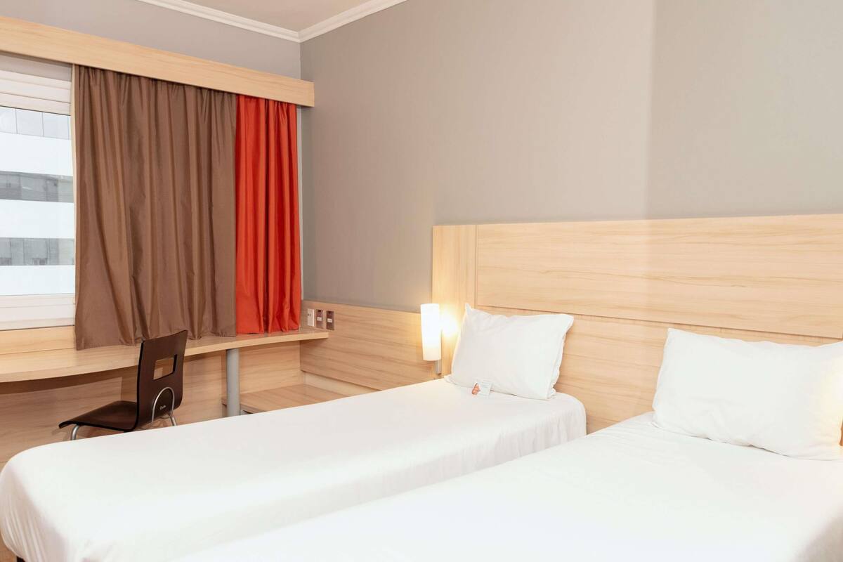 A hotel room with two single beds, a wooden headboard, a desk with a chair, and a window with curtains.