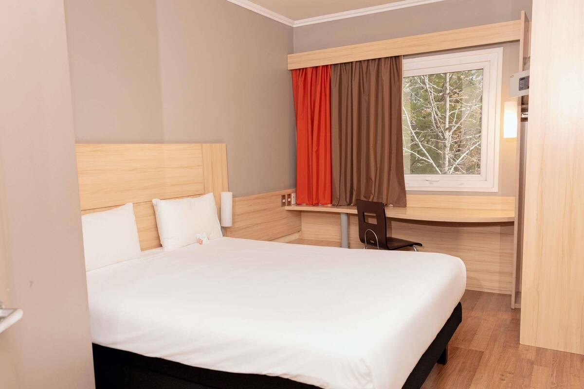 A hotel room with a bed, a desk, a chair, a window with curtains, and a view of trees.