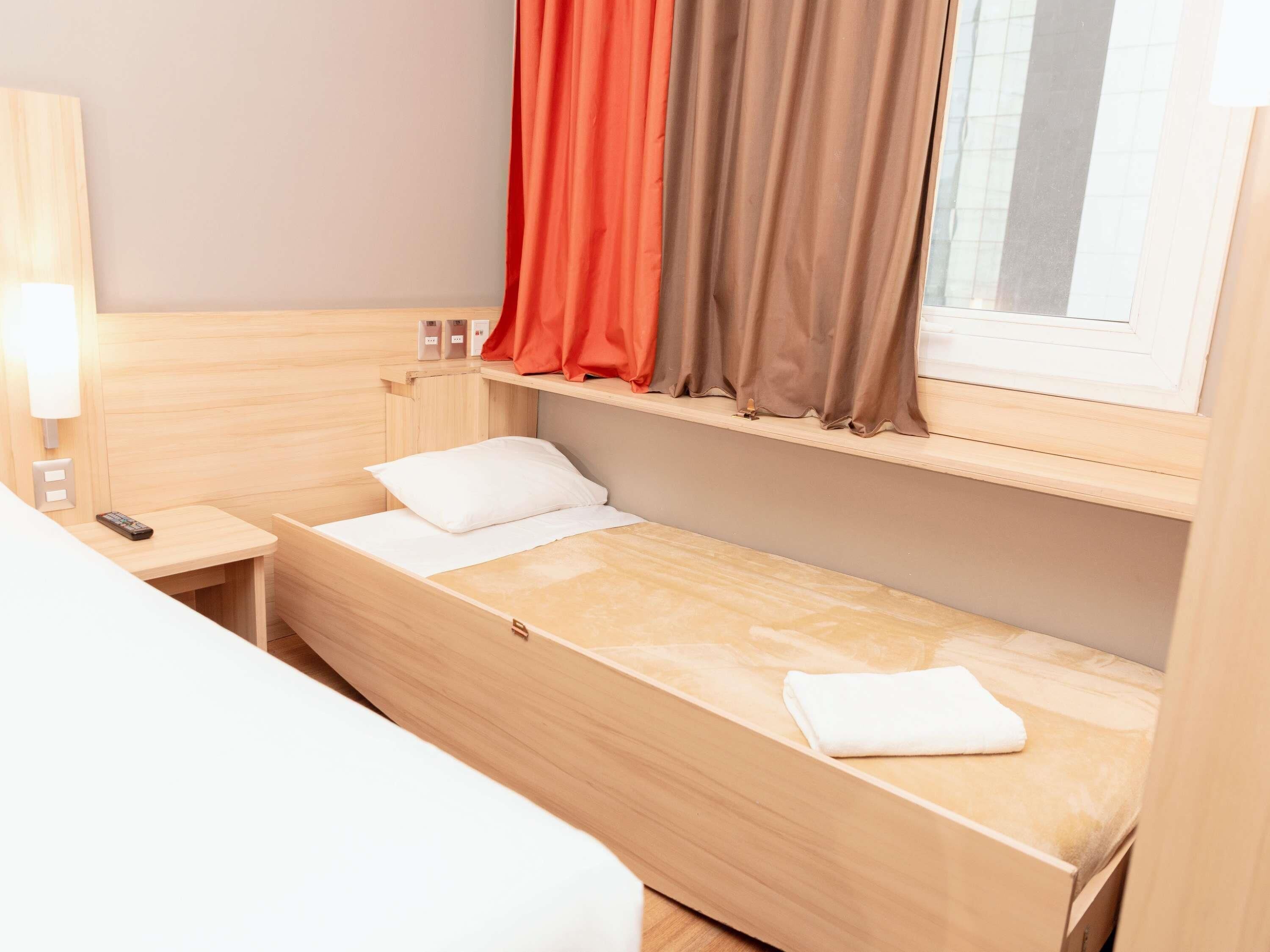 A small, single-bed room with a wooden headboard, a bedside table with a remote control, and a window with curtains.