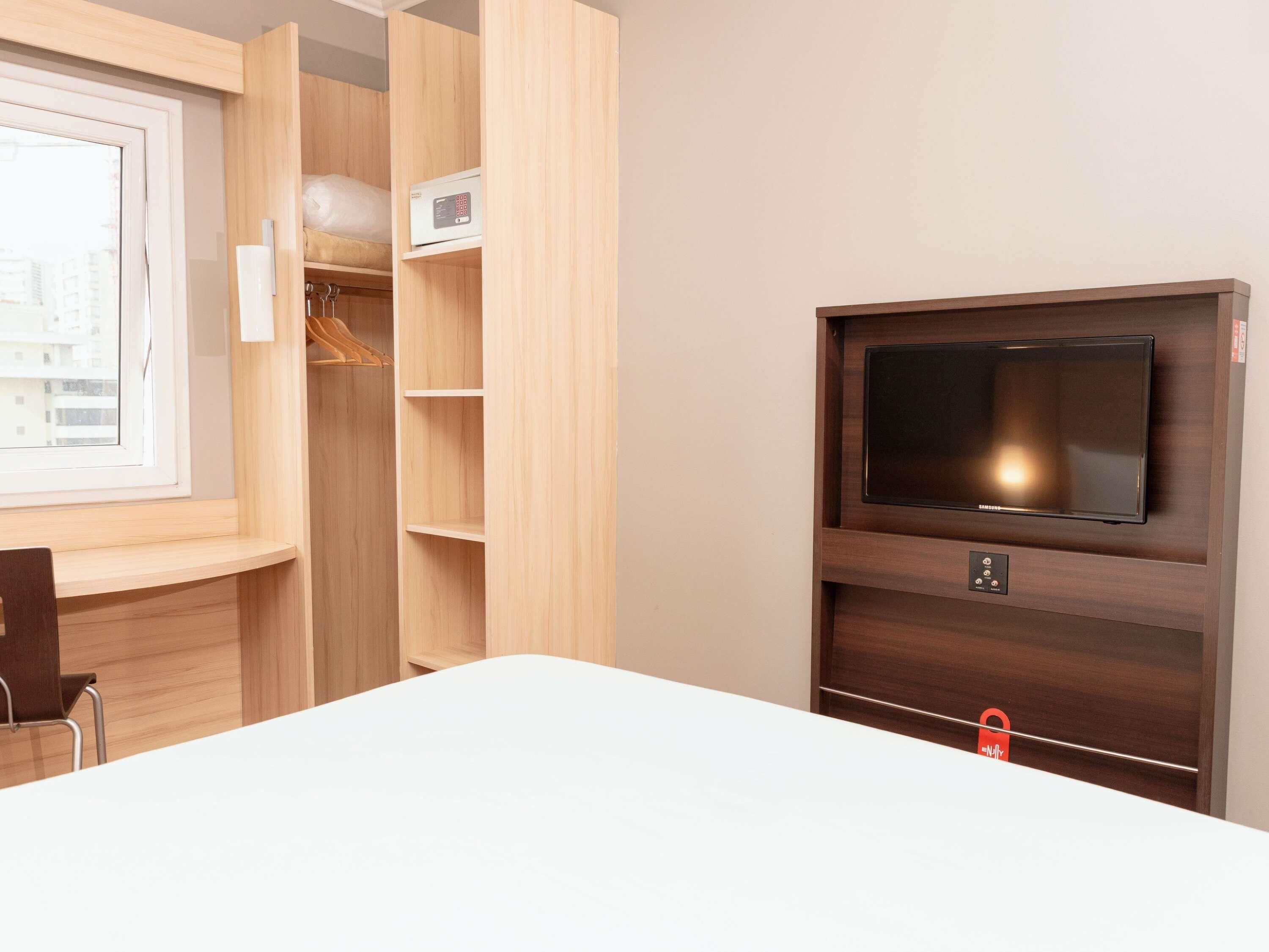 A hotel room with a bed, a TV mounted on the wall, wooden shelving, and a window.