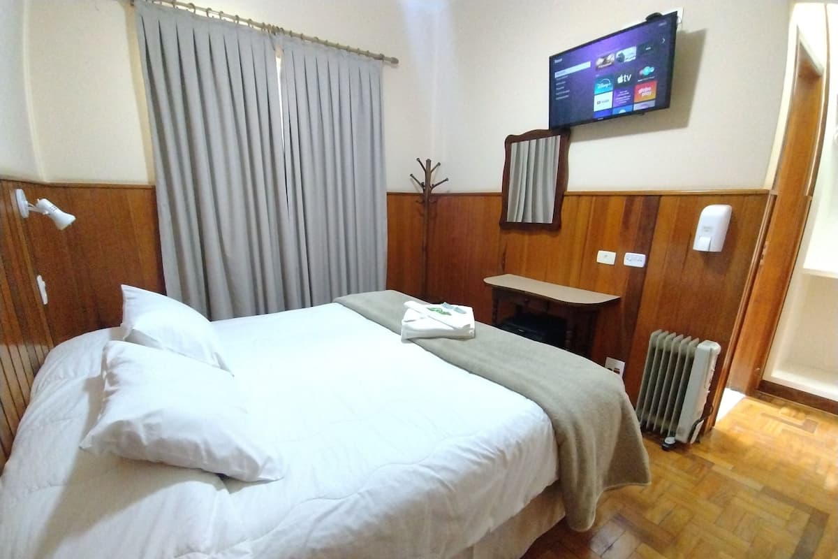 Habitación de hotel con cama, televisor montado en la pared, cabecero de madera, una mesita pequeña y un radiador.