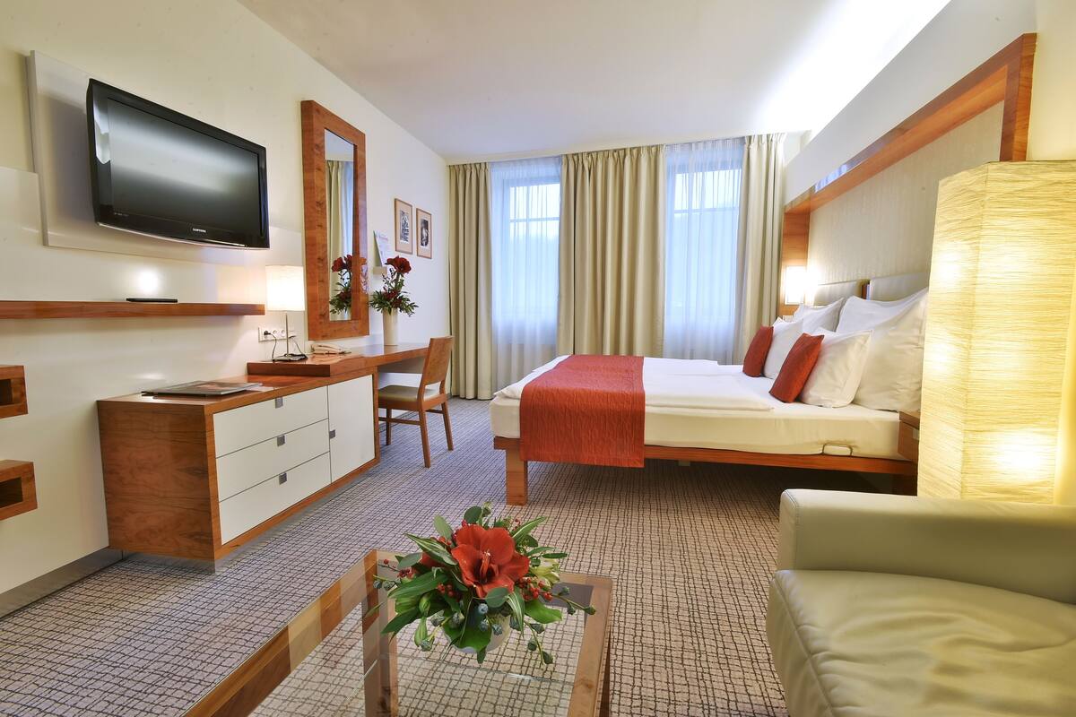 A hotel room with a bed, a desk, a TV, and a flower arrangement.