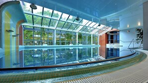 A modern indoor swimming pool with a glass ceiling, a curved seating area, and a view of greenery outside.
