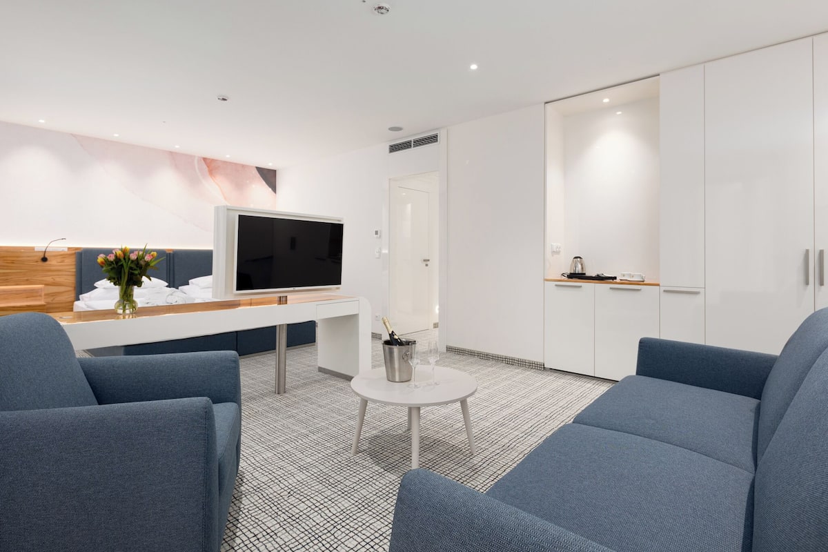 A modern hotel room with a flat-screen TV, a small table with a vase and glasses, and a sofa.