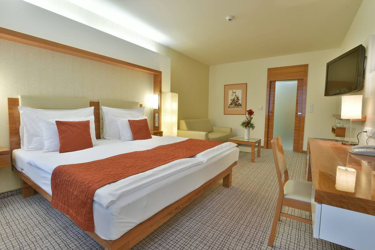 A hotel room with a large bed, a desk, a chair, a TV, and a small table with a vase of flowers.