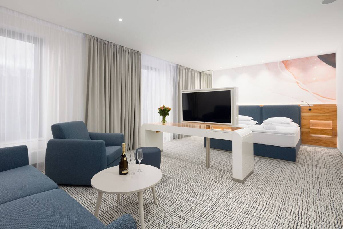A modern hotel room with a large flat-screen TV, a sofa, a small table with a bottle and glass, and a bed with a headboard.