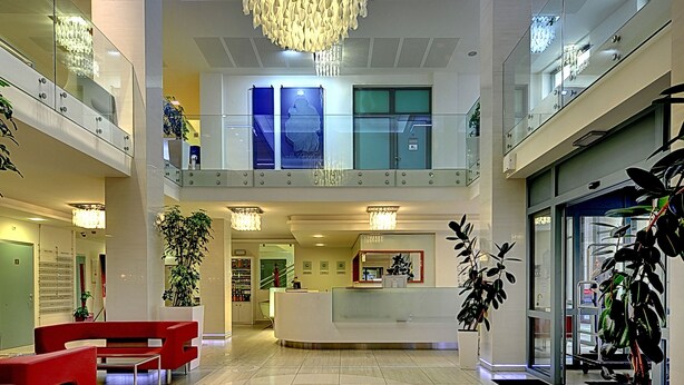 A spacious lobby with a central reception desk, red seating, and a modern chandelier.