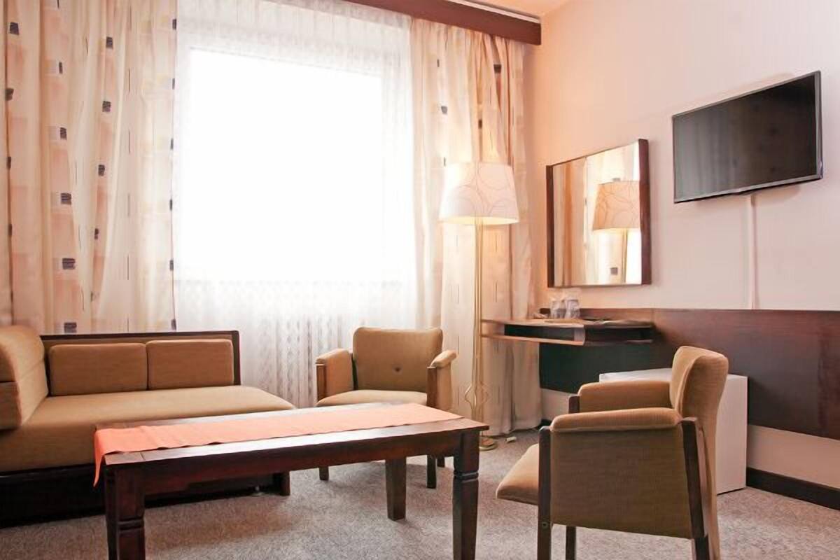A hotel room with a sofa, two armchairs, a wooden coffee table, a desk with a lamp, a flat-screen TV, and a large window with curtains.
