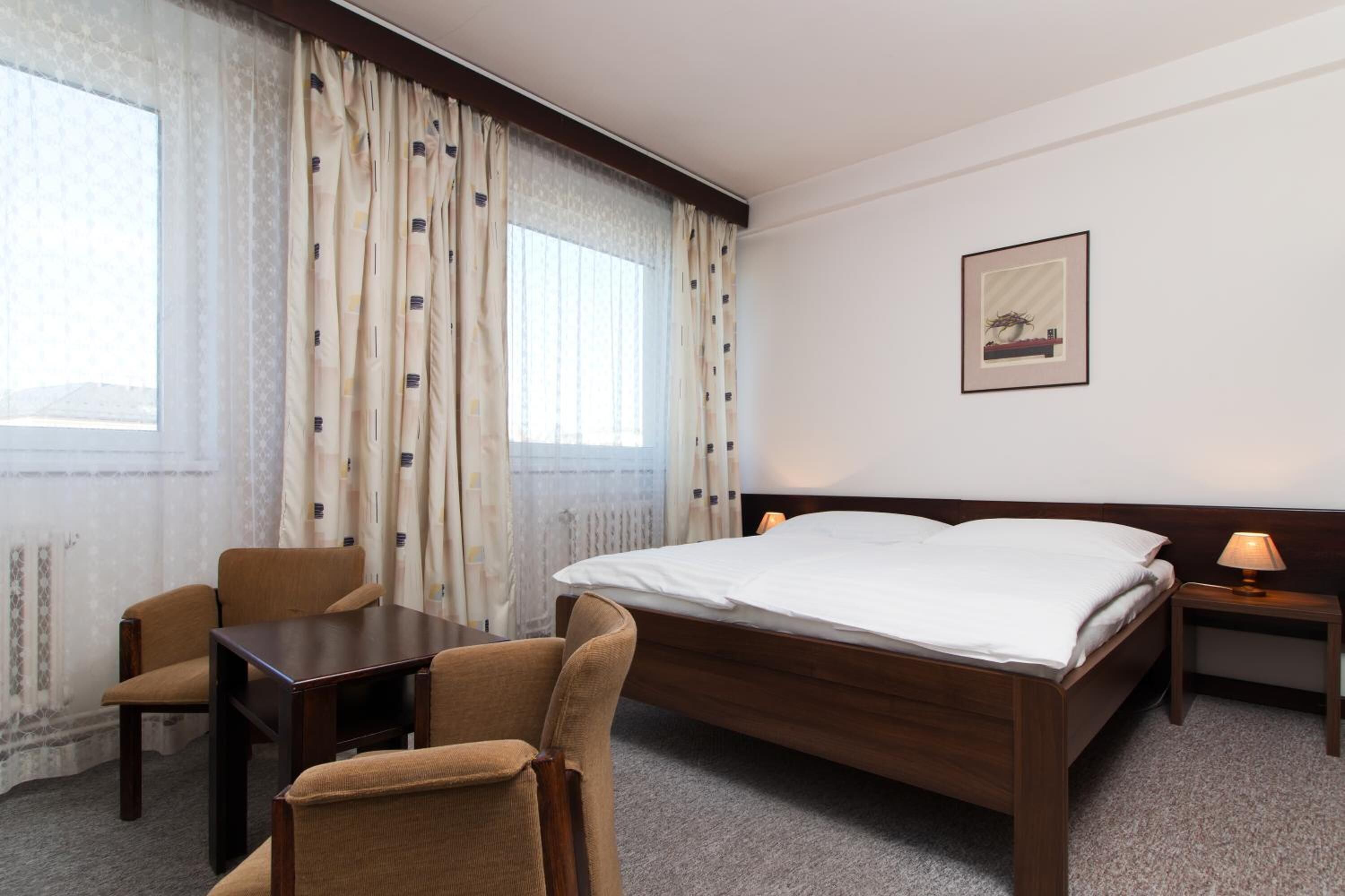 A hotel room with a large bed, a desk, two chairs, a nightstand with a lamp, and a window with curtains.