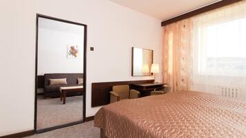 A hotel room with a large bed, a desk with a chair, a sofa, a mirror, and a window with curtains.