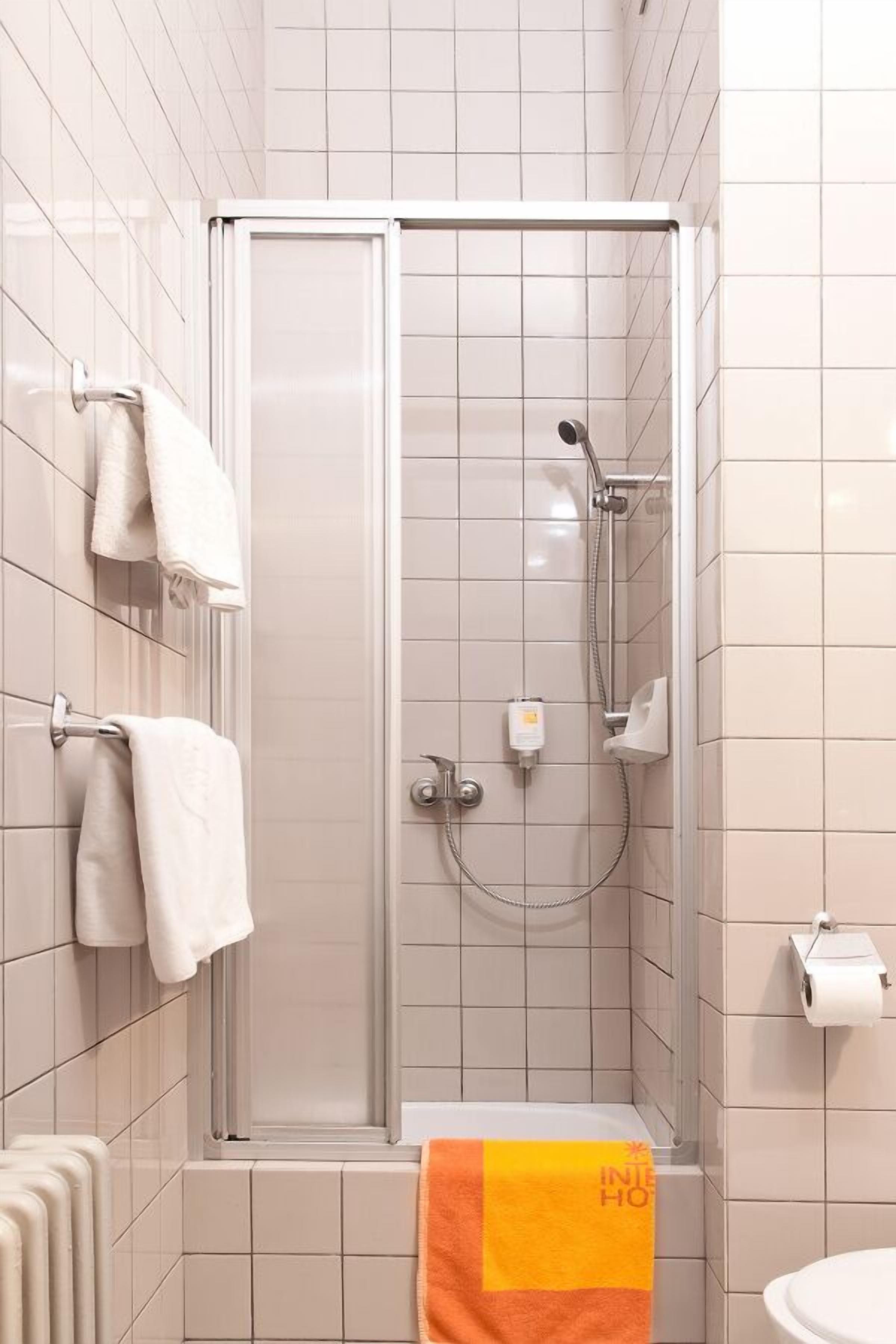 A modern bathroom with a glass-enclosed shower, white tiles, a towel rack with white towels, and a single orange towel on the floor.
