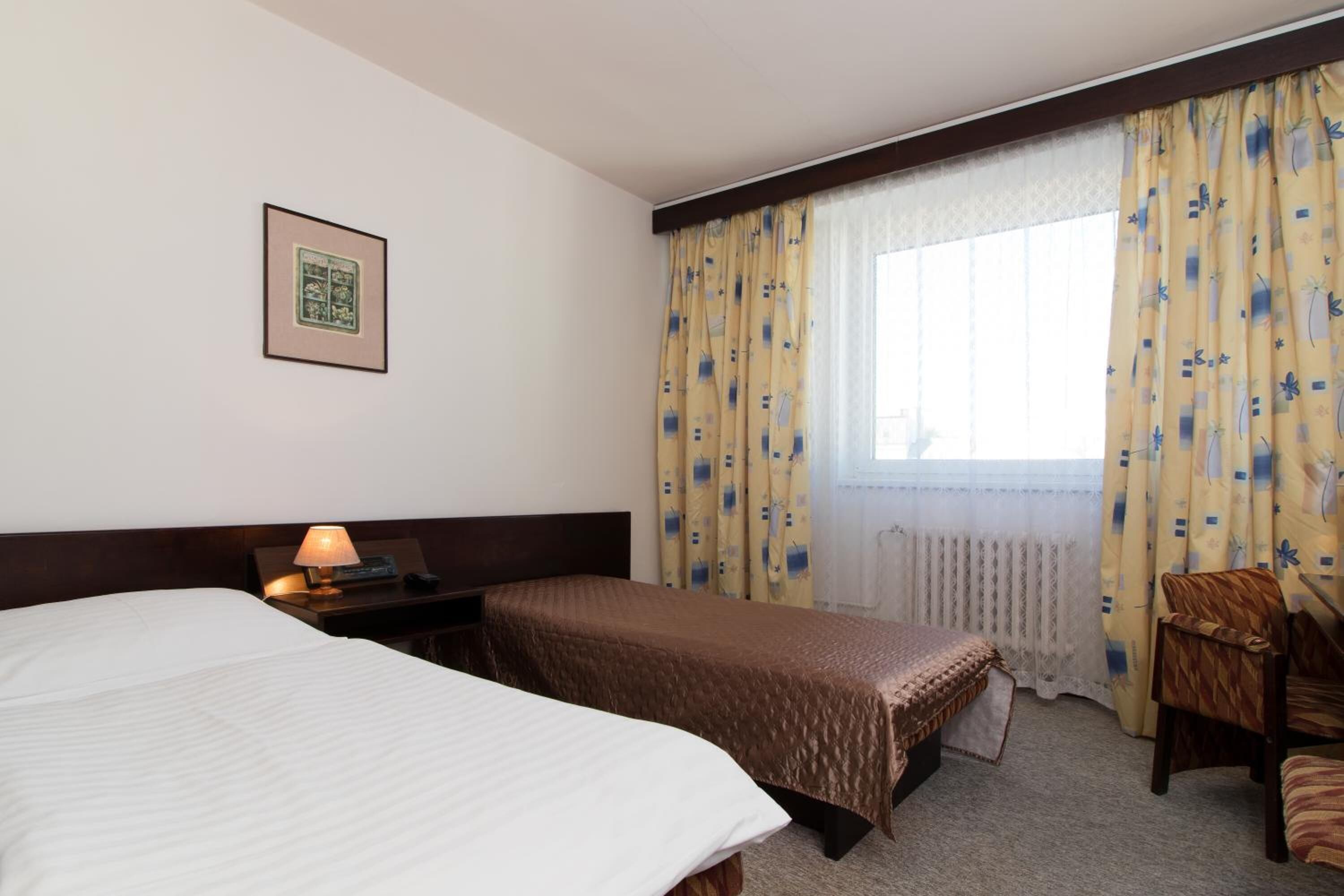 A hotel room with two beds, a nightstand with a lamp, a chair, and a window with curtains.