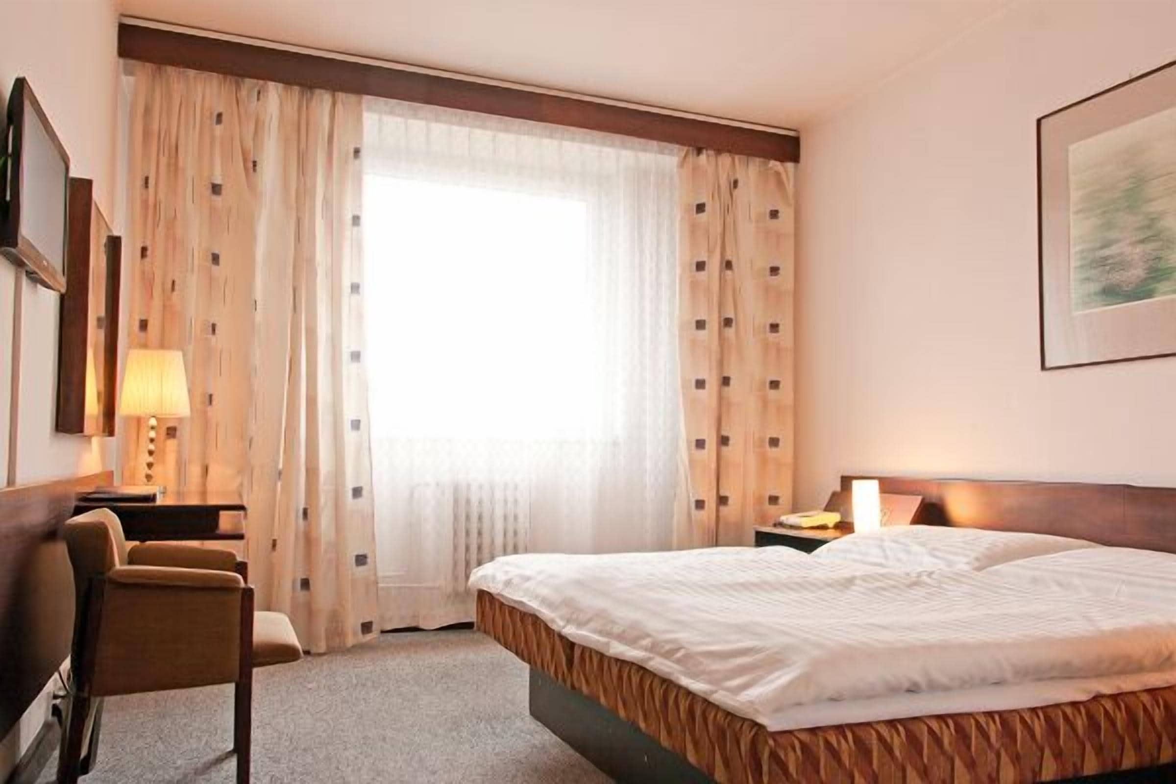 Une chambre d’hôtel avec un lit, une chaise, un bureau et une fenêtre avec des rideaux.