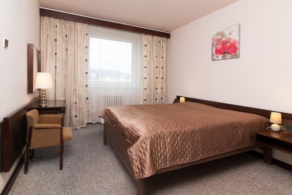 A hotel room with a bed, a chair, a desk, a lamp, and a painting on the wall.