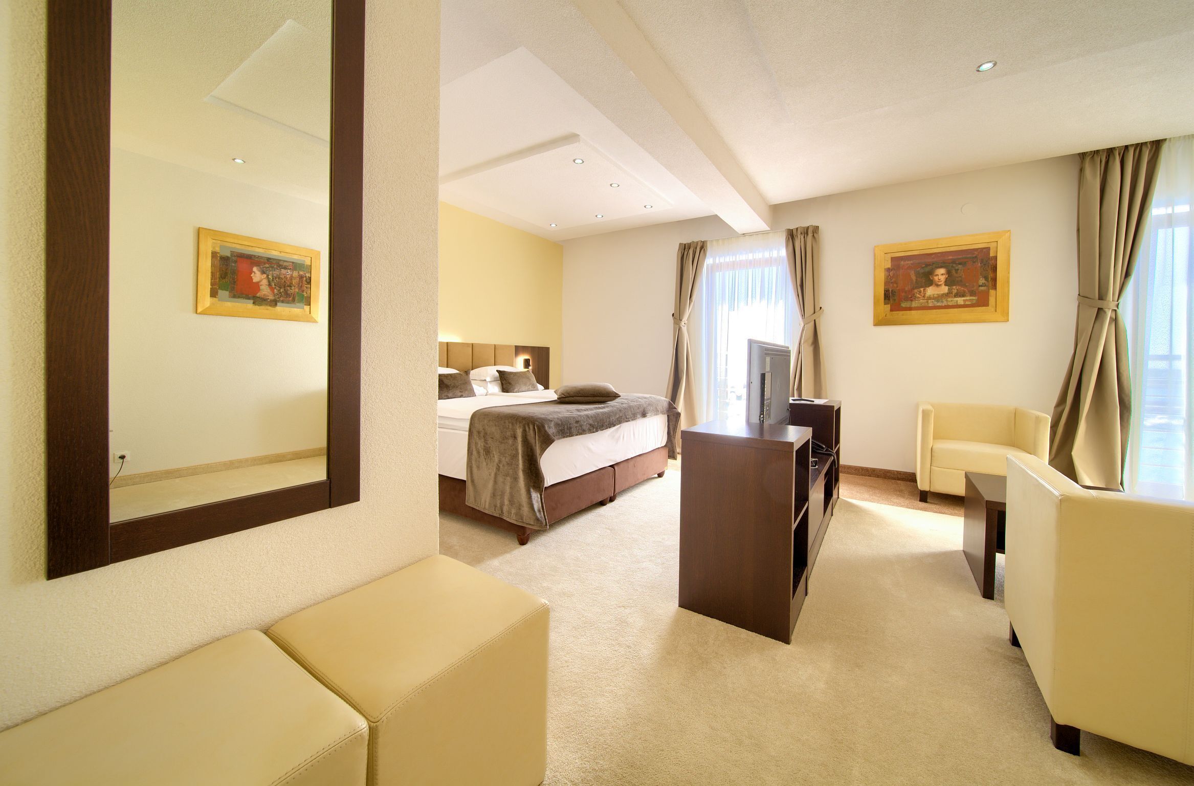 A hotel room with a bed, a desk, a chair, a sofa, a mirror, and two paintings on the walls.