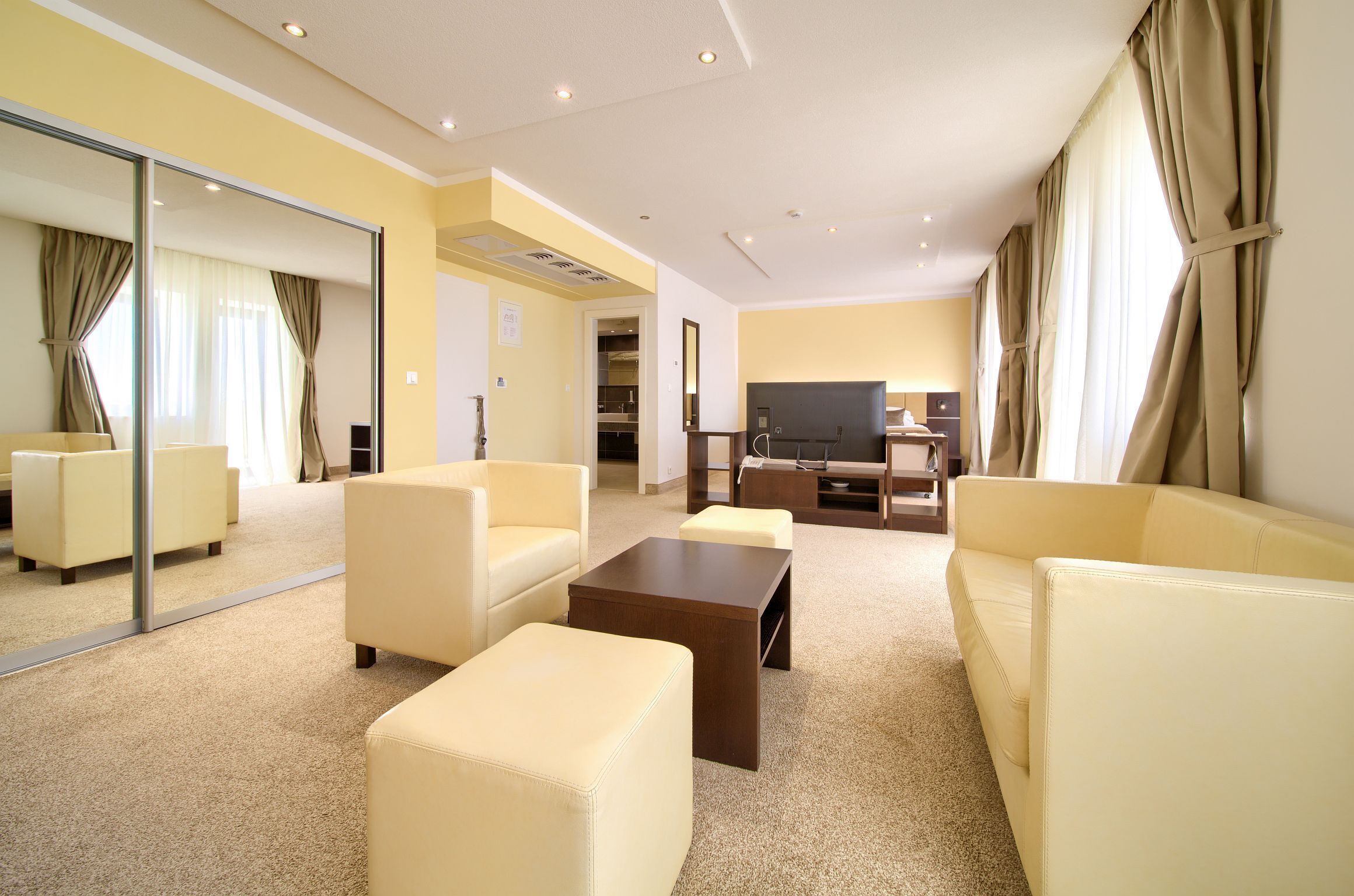 A modern hotel room with a large mirror, a flat-screen TV, and a seating area with sofas and ottomans.