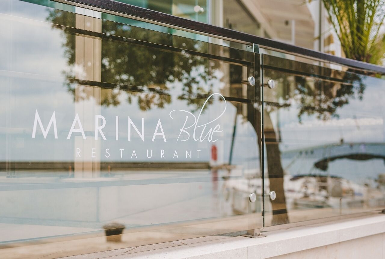 A glass window with the text "MARINA Blue RESTAURANT" and reflections of trees and boats.