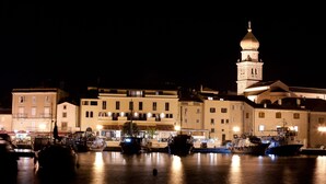 Un puerto deportivo frente al agua de noche, con varios barcos amarrados, un edificio destacado con una torre y ventanas iluminadas.