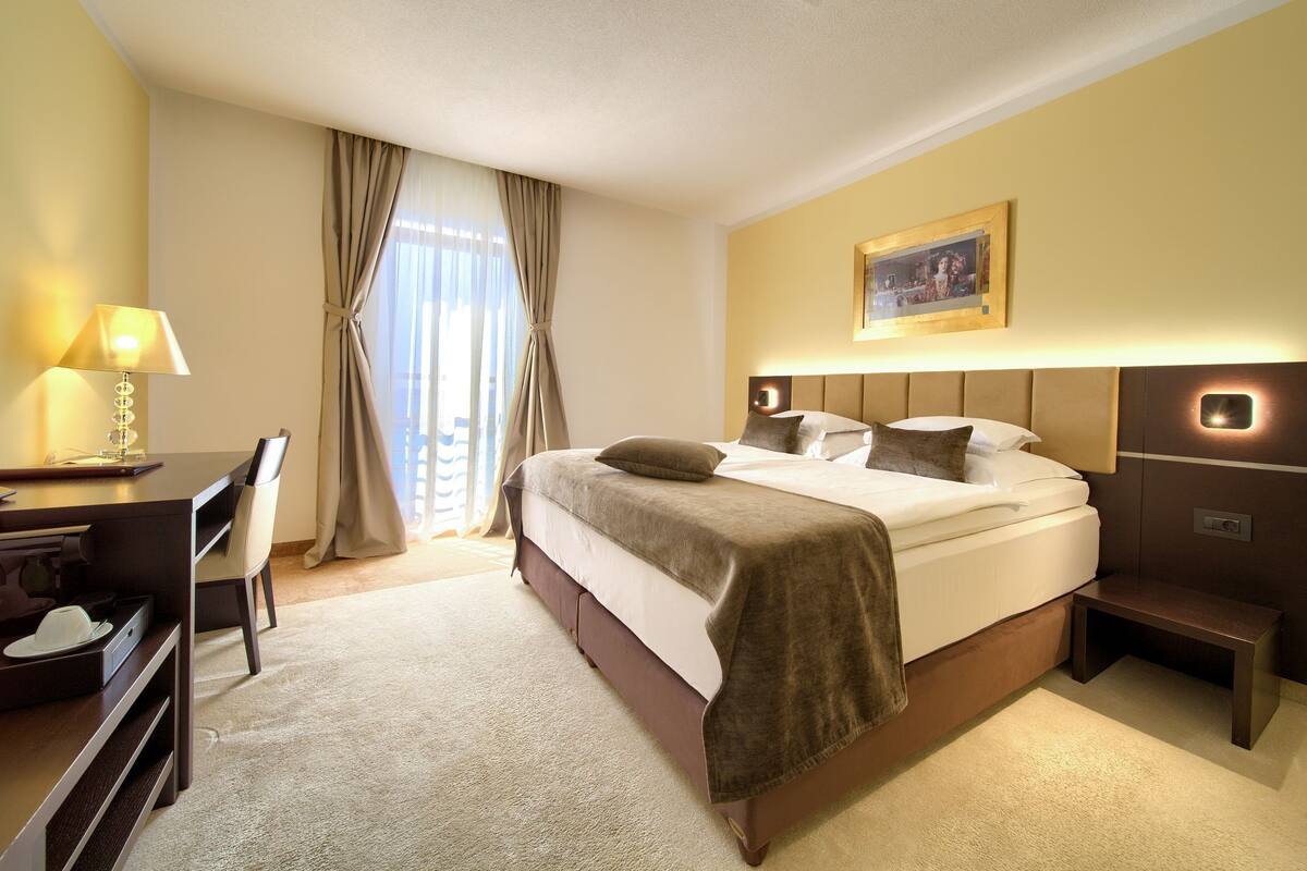 Een hotelkamer met een bed, bureau, stoel, lamp en een schilderij aan de muur.