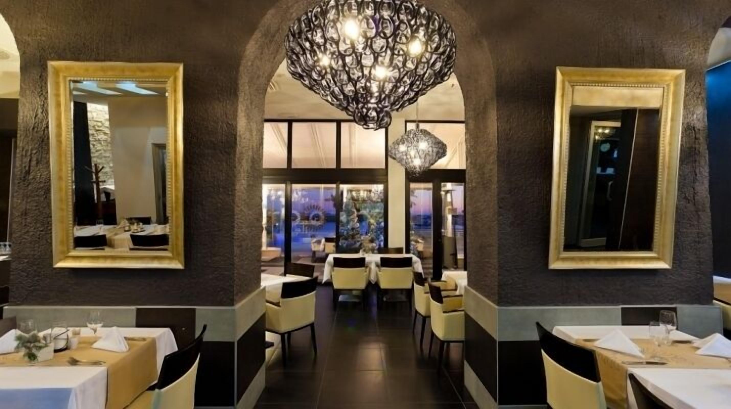 A restaurant with a central dining area, framed by large mirrors and a decorative chandelier. Tables are set with white tablecloths and chairs.