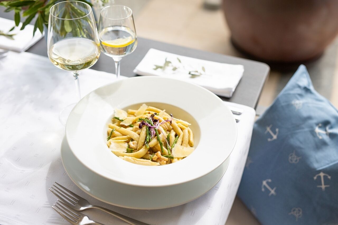 A dining table with a plate of pasta, two glasses of white wine, and a blue napkin with anchor patterns.