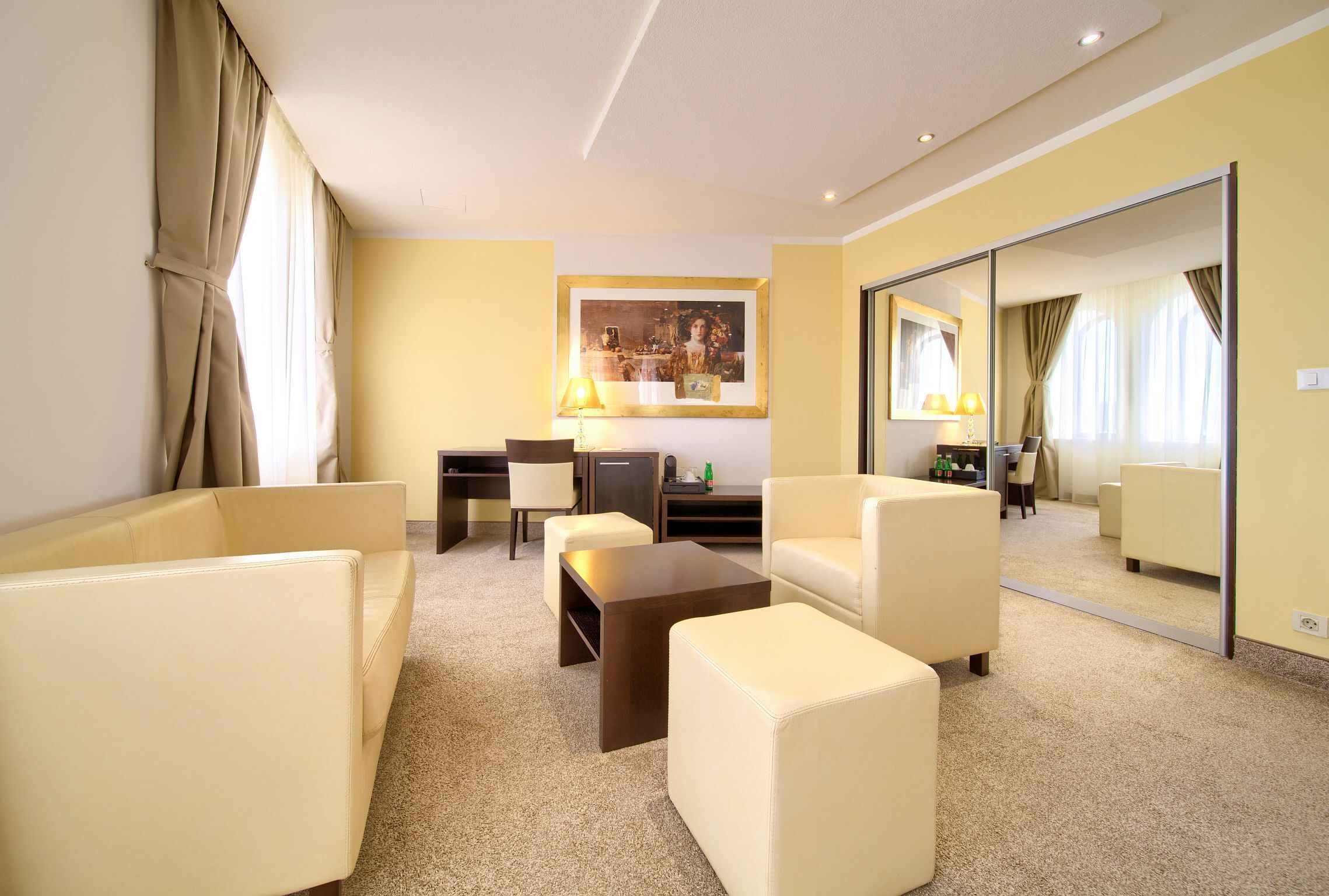 Ein Hotelzimmer mit Sofa, Ohrensessel, Couchtisch, Schreibtisch, Lampe und einem großen Spiegel.