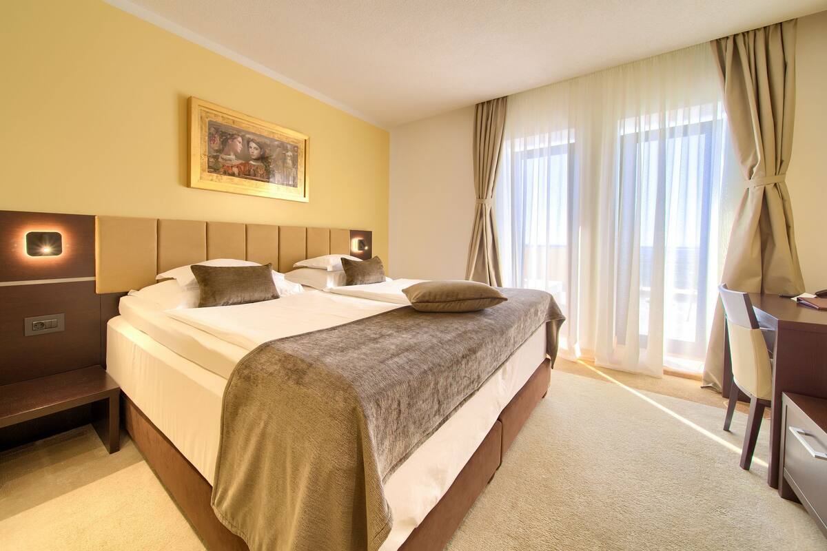 Hotelkamer met een groot bed, een bureau en een stoel. Er hangt een schilderij aan de muur en er is een raam met gordijnen.