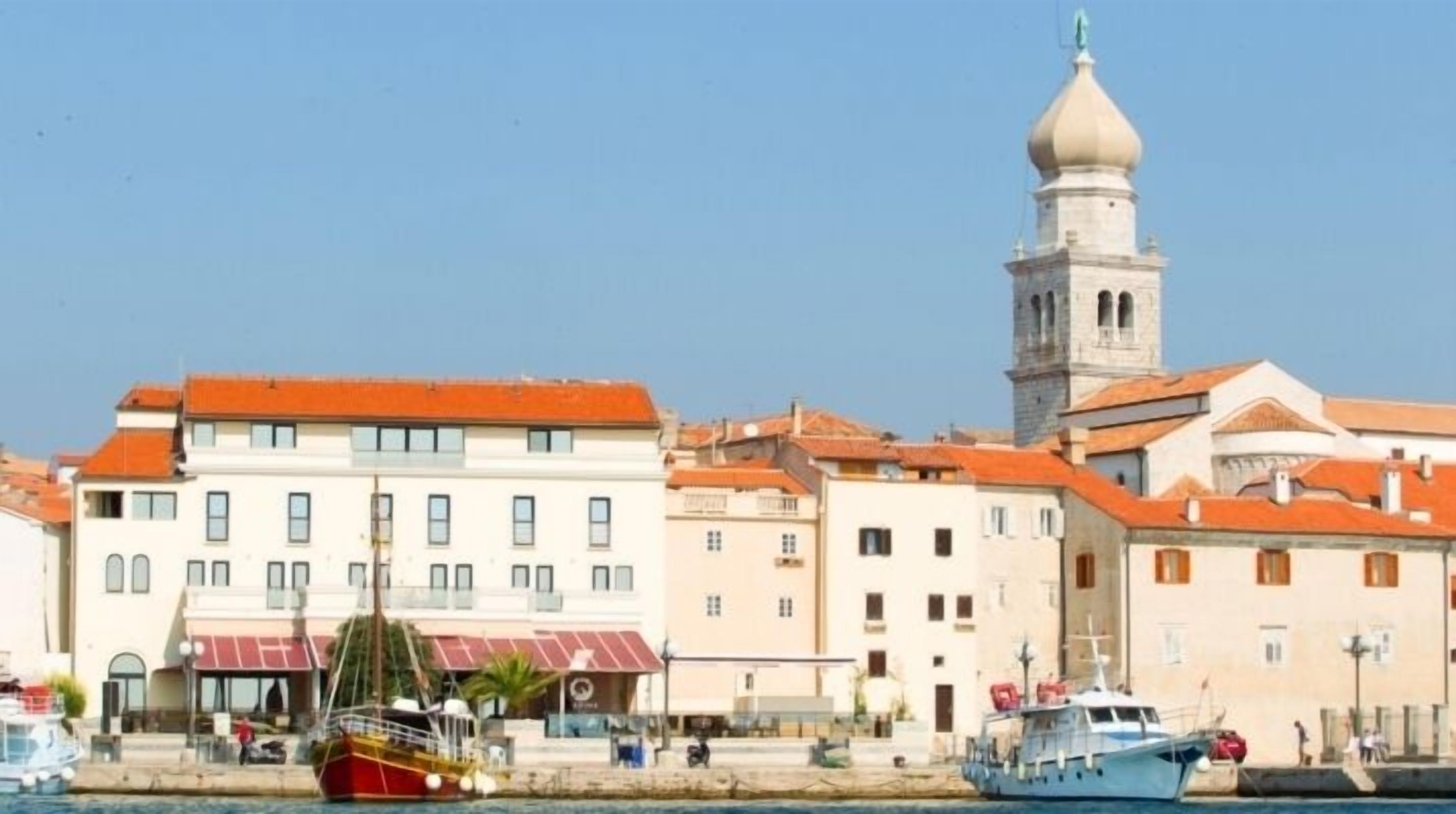 Une ville côtière qui se distingue par une tour imposante, un bâtiment rouge et blanc doté d’un balcon, et un bateau bleu amarré au quai.