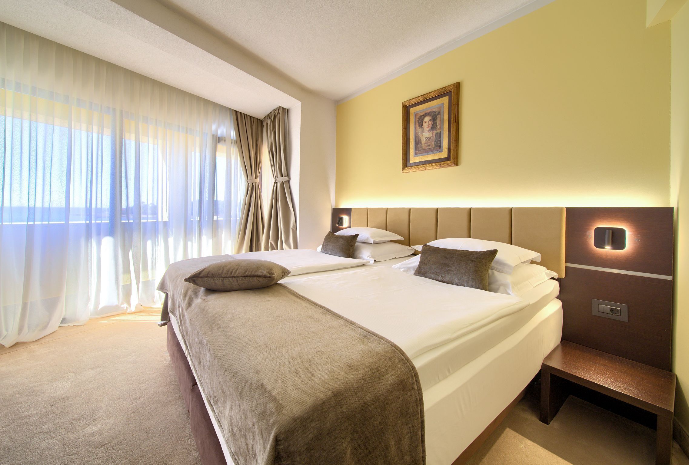 Quarto de hotel com duas camas, uma mesa de cabeceira, um criado-mudo, um quadro na parede e uma janela com cortinas leves.