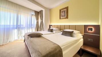 Quarto de hotel com duas camas, uma mesa de cabeceira, um criado-mudo, um quadro na parede e uma janela com cortinas leves.