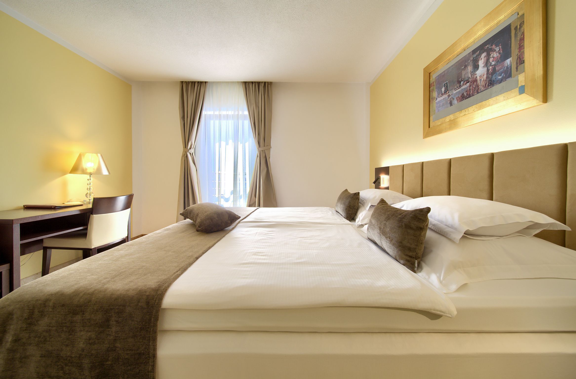 A hotel room with a bed, a desk, a chair, and a painting on the wall.