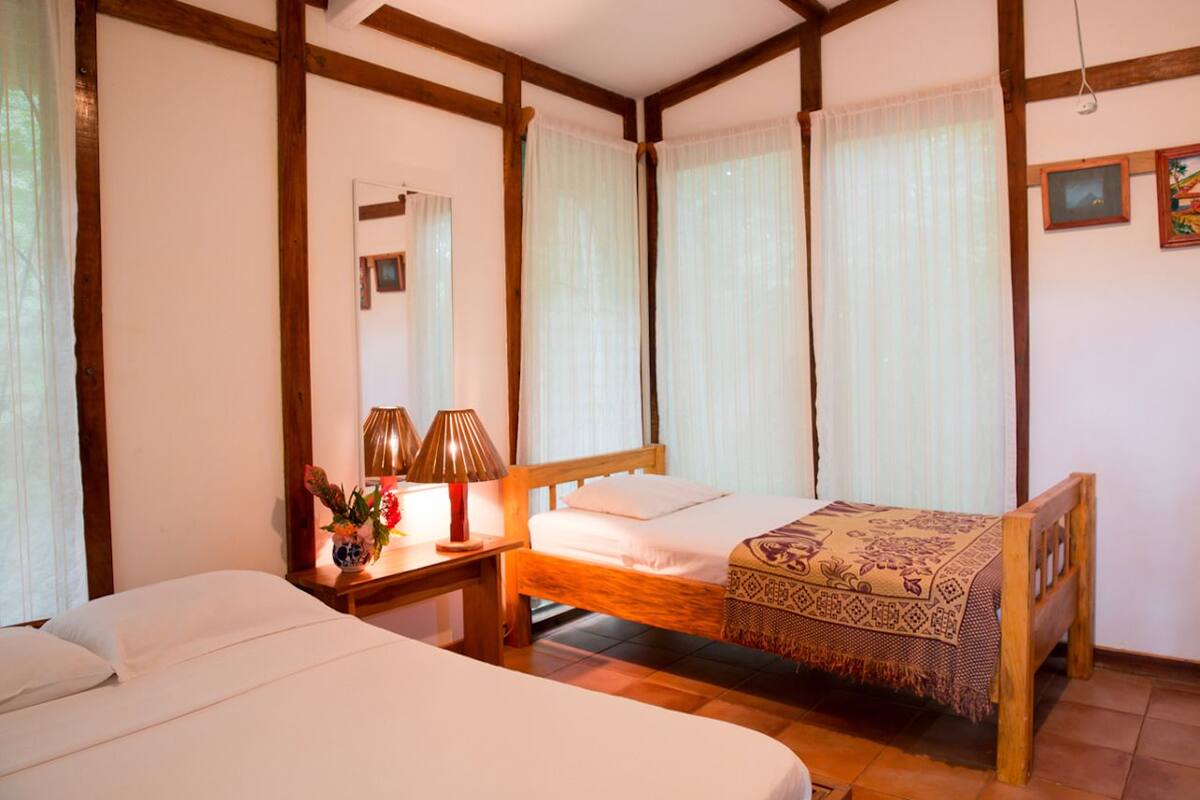A room with two beds, wooden beams, a mirror, a bedside table with lamps, and sheer curtains.