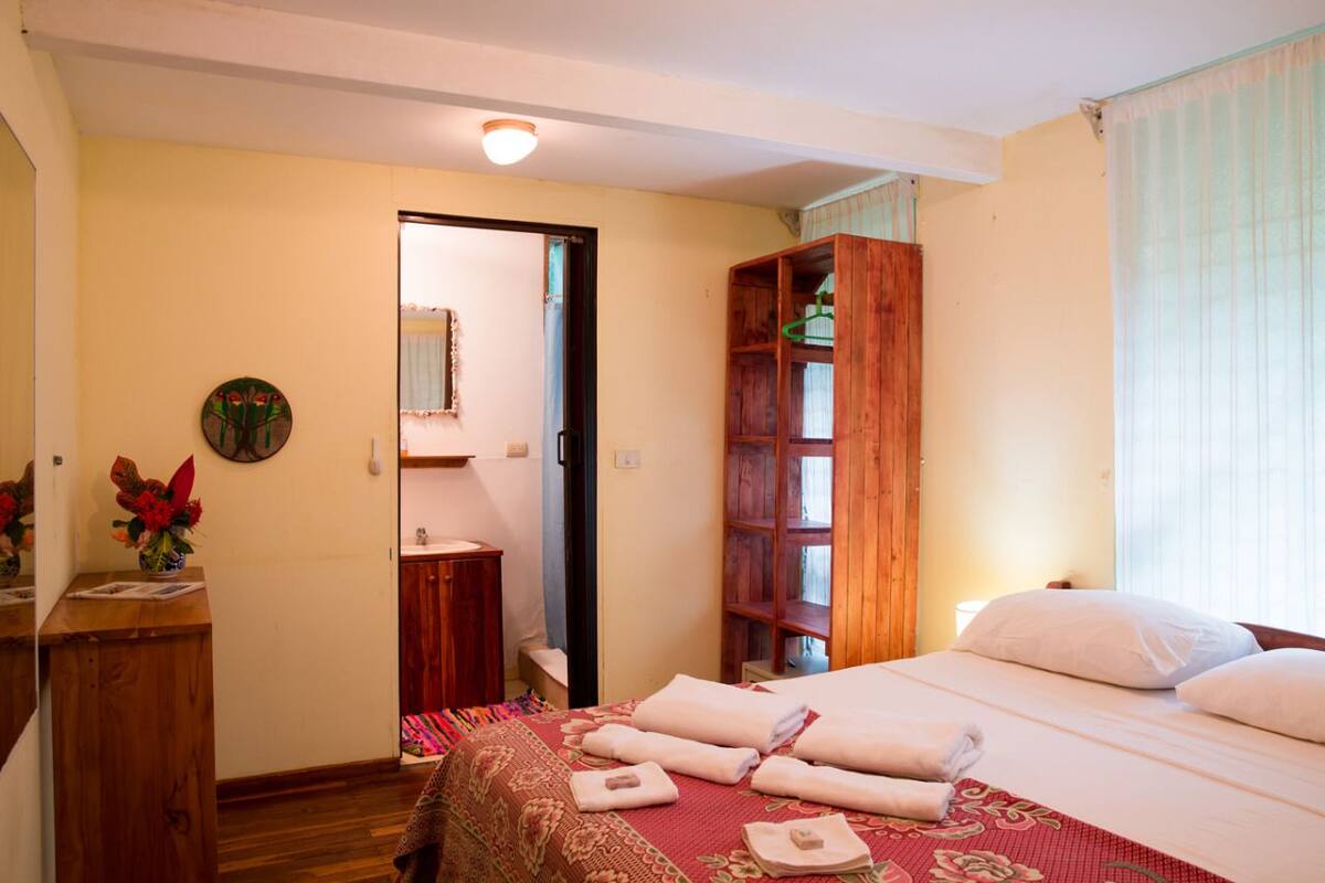 A hotel room with a bed, a wooden wardrobe, a desk with a vase of flowers, and a door leading to another room.