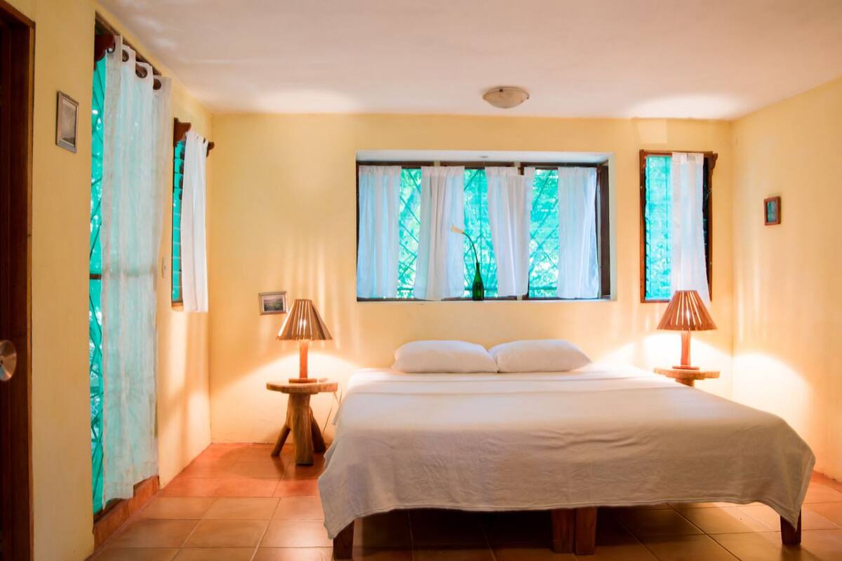 A bedroom with a large bed, two bedside tables with lamps, a window with curtains, and a door.