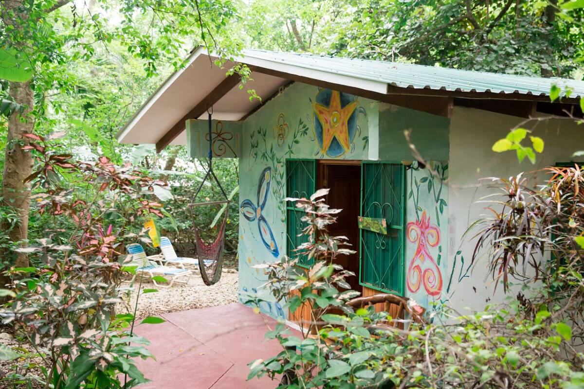 A small, rustic hut with a sloped roof and colorful murals, surrounded by lush greenery and a hammock.