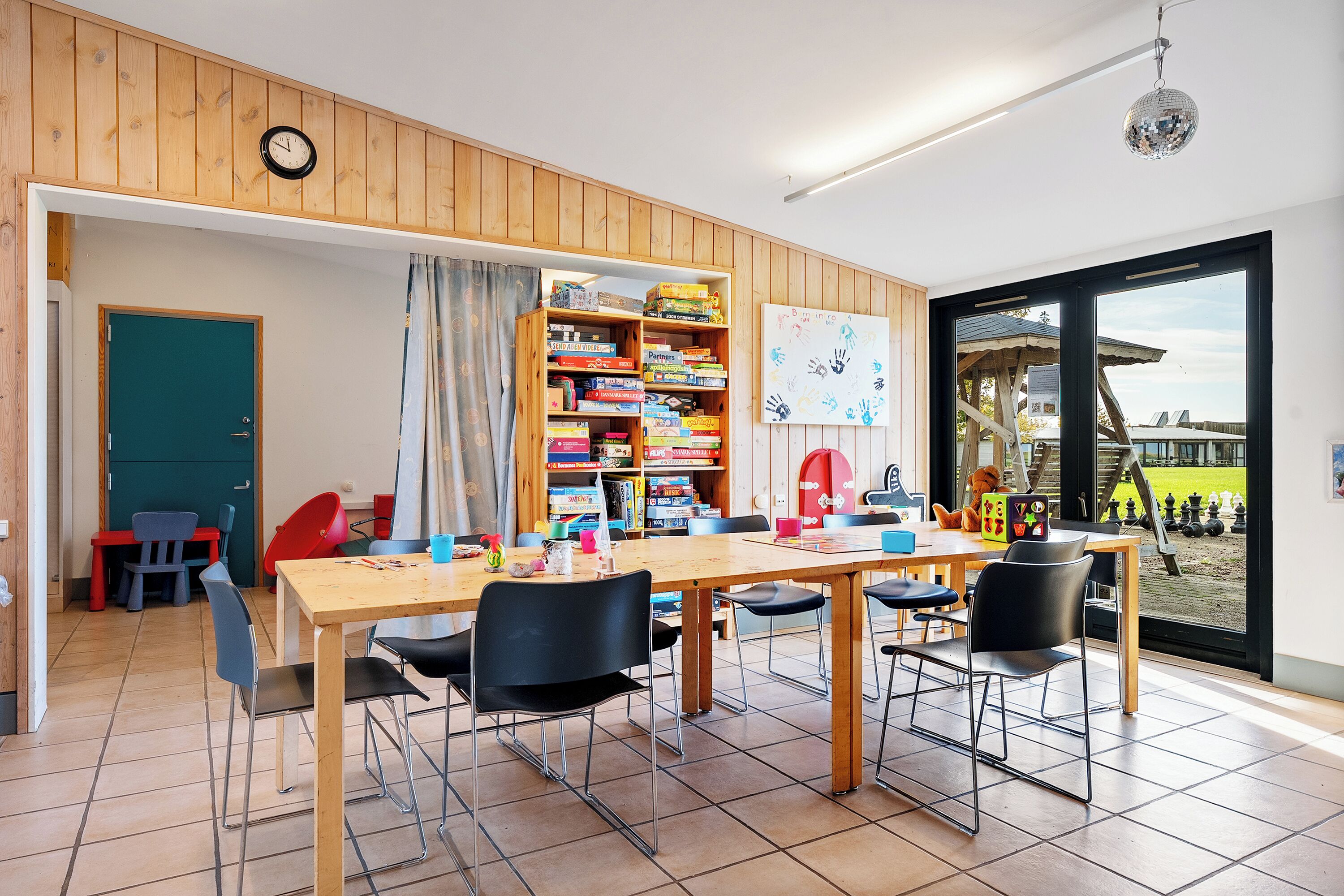 A room with a wooden table, chairs, a bookshelf, a clock, and a view of the outside.