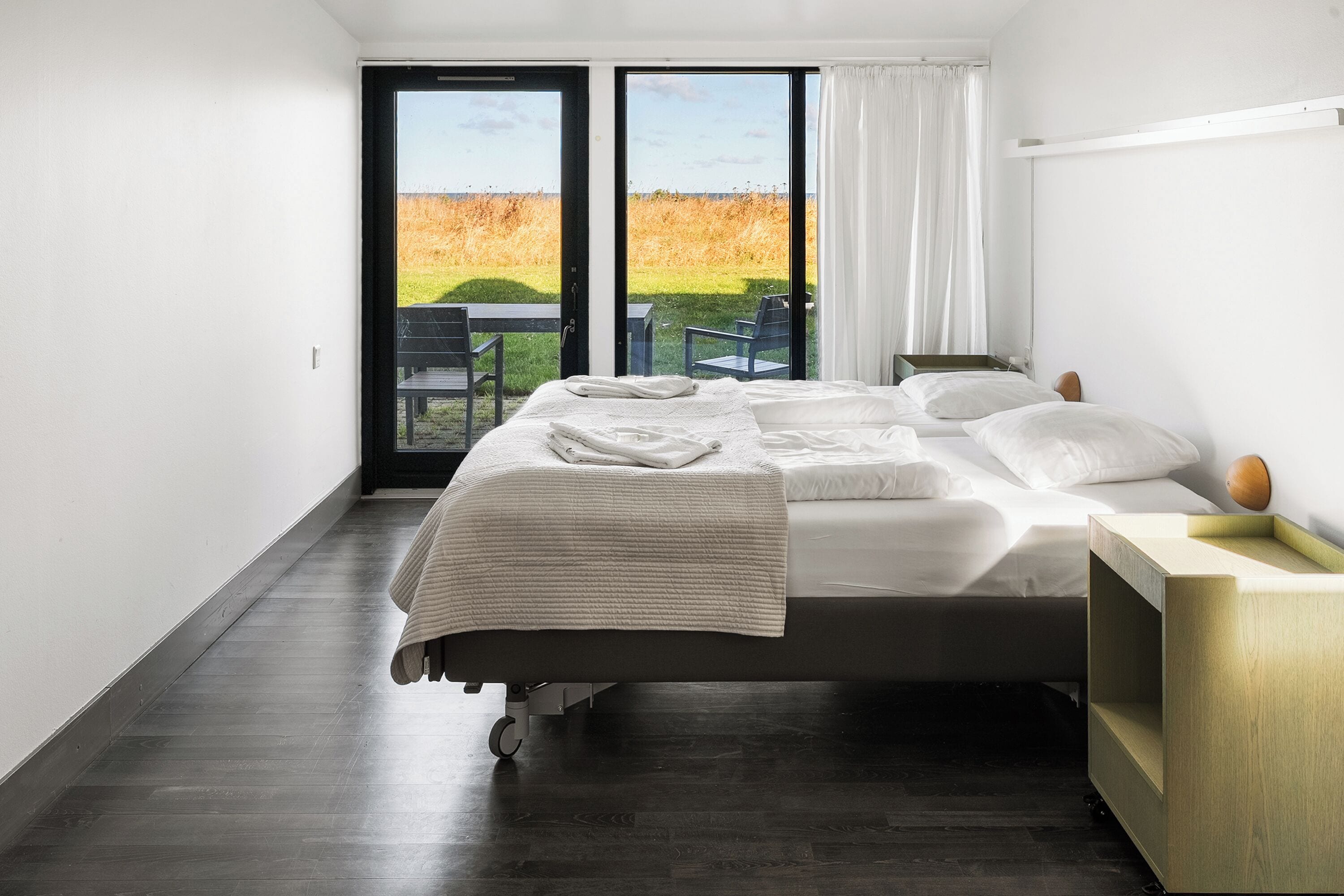 Un dormitorio moderno con una cama grande, una mesita de noche de madera y vistas a un campo cubierto de hierba a través de una puerta corredera.