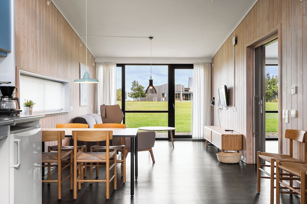 Une cuisine moderne avec un lambris en bois, un coin repas avec une table et des chaises, et une vue sur l’extérieur grâce à de grandes portes vitrées.