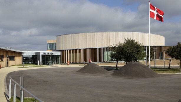 A modern building with a circular design, a flagpole with a flag, and a paved area with mounds. - Musholm Ferie-Sport-Konference (Korsor)