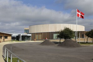 Ein modernes Gebäude mit kreisförmigem Design, einer Fahnenmast mit einer Flagge und einer gepflasterten Fläche mit Erhöhungen.