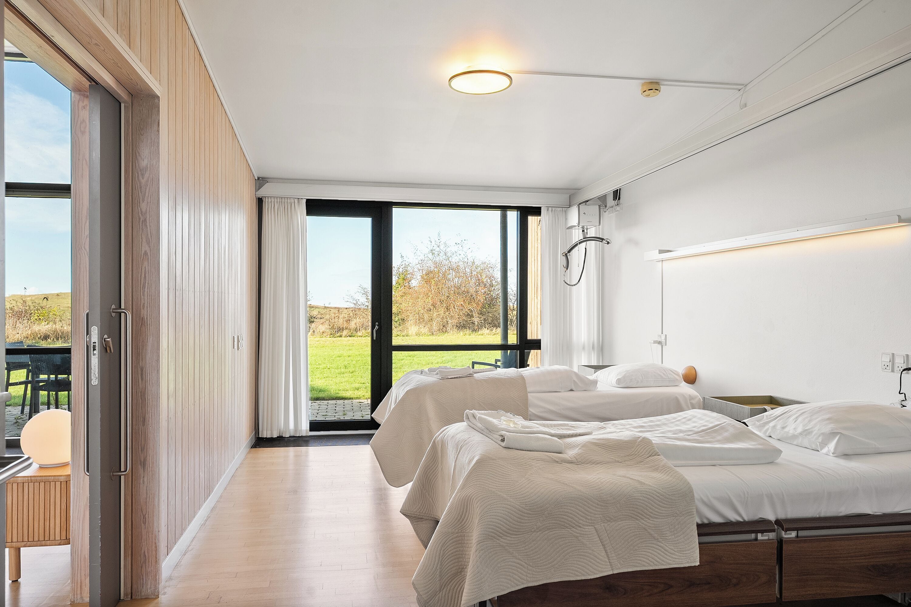 Une chambre d’hôtel moderne dotée d’une grande porte-fenêtre coulissante donnant sur un espace extérieur, d’un lit avec une literie blanche et d’une table de chevet.