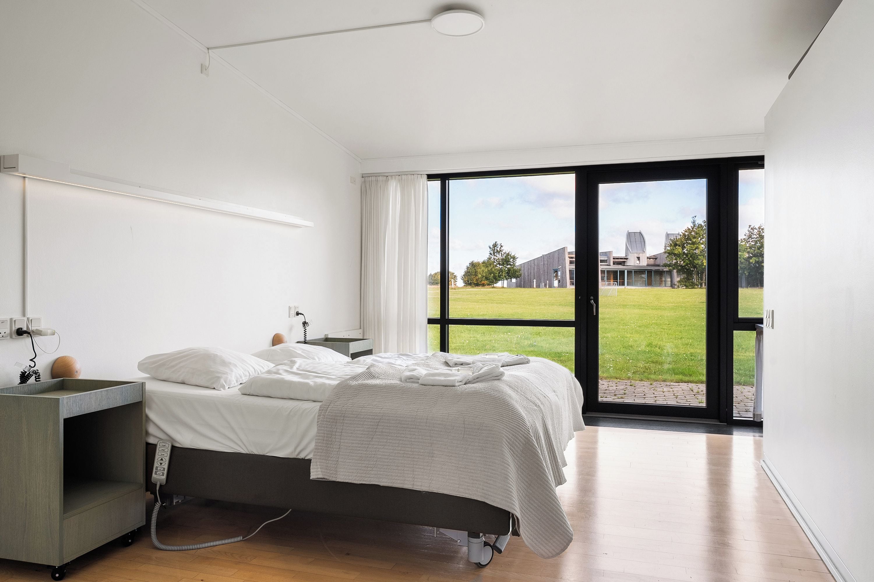 A modern bedroom with a bed, bedside table, and a view of a grassy area through a large window.