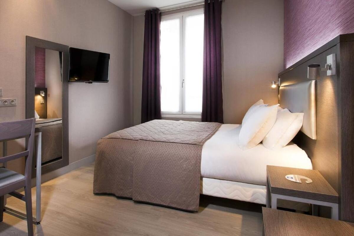 A modern hotel room with a bed, a TV, a desk, and a window with curtains.