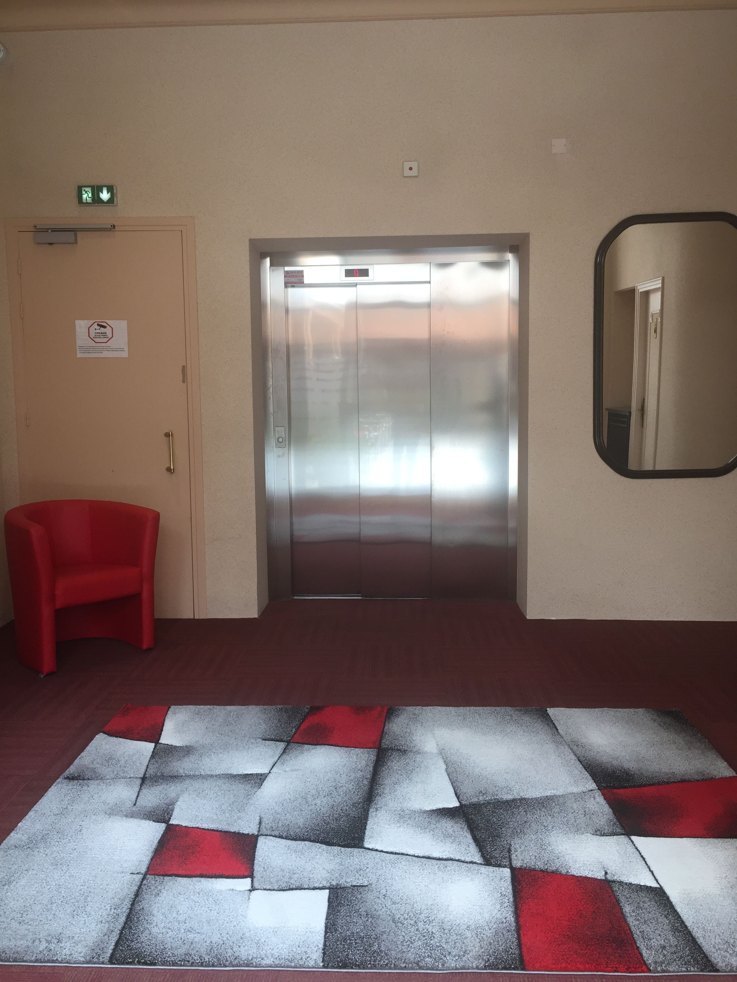 Un ascensor con pantalla digital, una silla roja y una alfombra de estampado geométrico.