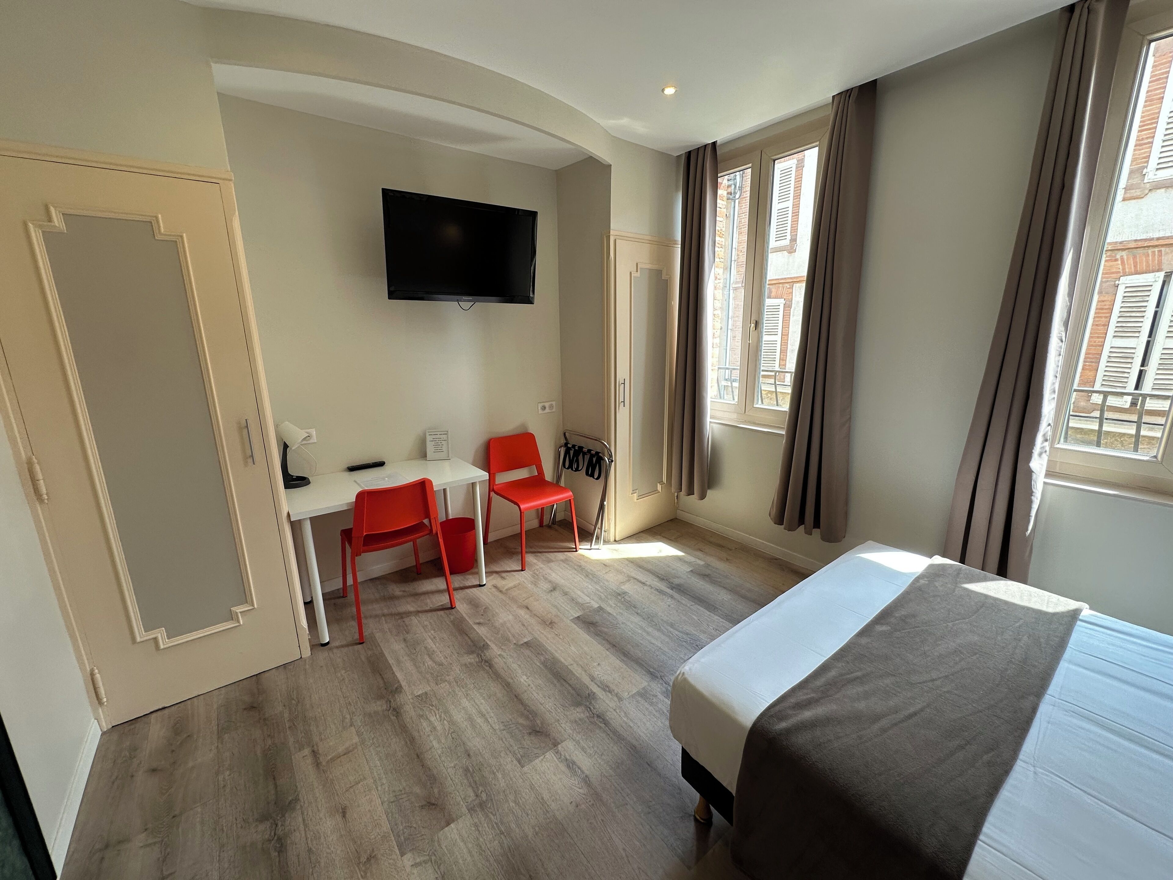 A hotel room with a bed, a desk with two red chairs, a television mounted on the wall, and large windows with curtains.