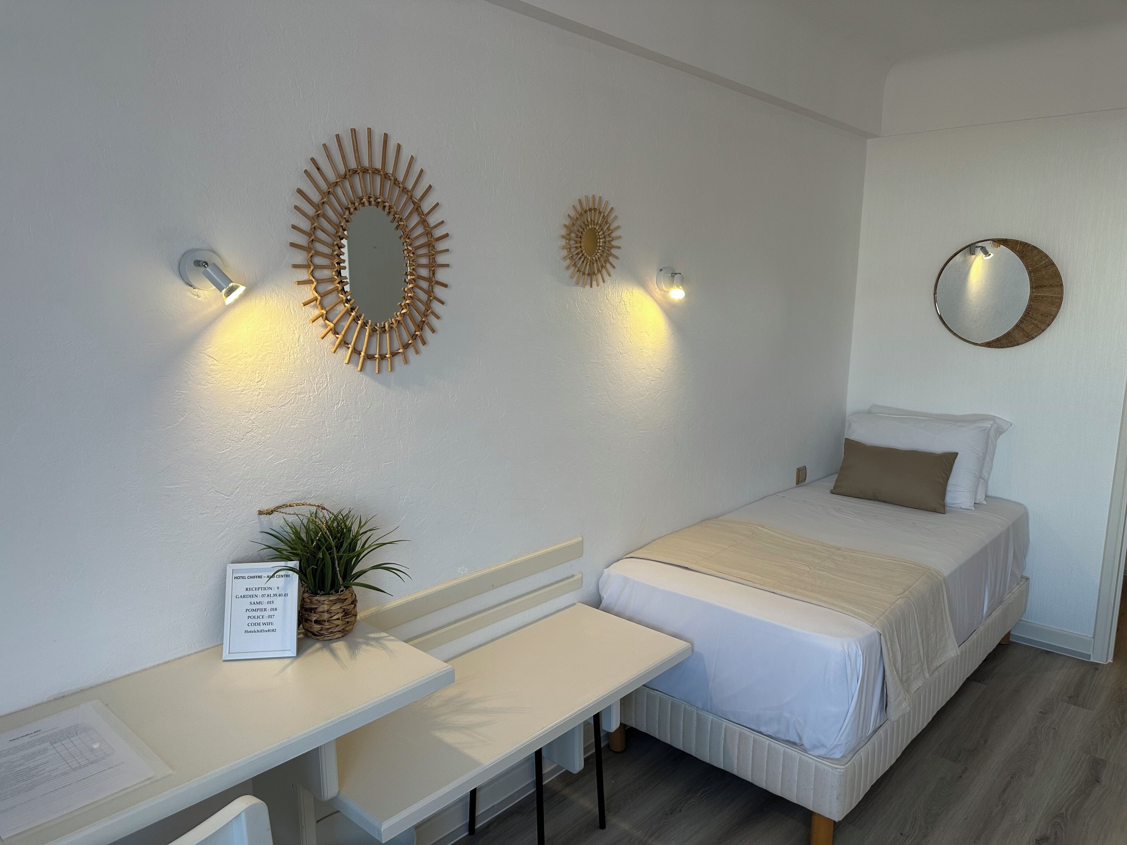 A hotel room with a bed, a desk, a chair, a small potted plant, and wall-mounted mirrors.
